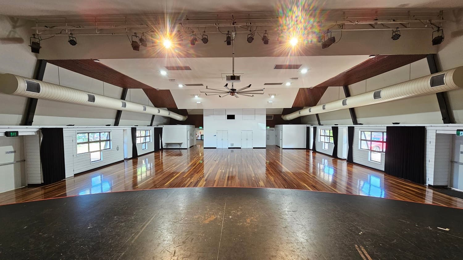 A spacious hall with polished wooden floors and a high ceiling. The ceiling features exposed beams, ductwork, and a large central fan. On the left and right walls, there are several windows with black curtains. The back wall has white doors and panels. Overhead, there are multiple lights and speakers. The foreground shows a slightly worn stage area with a dark surface.