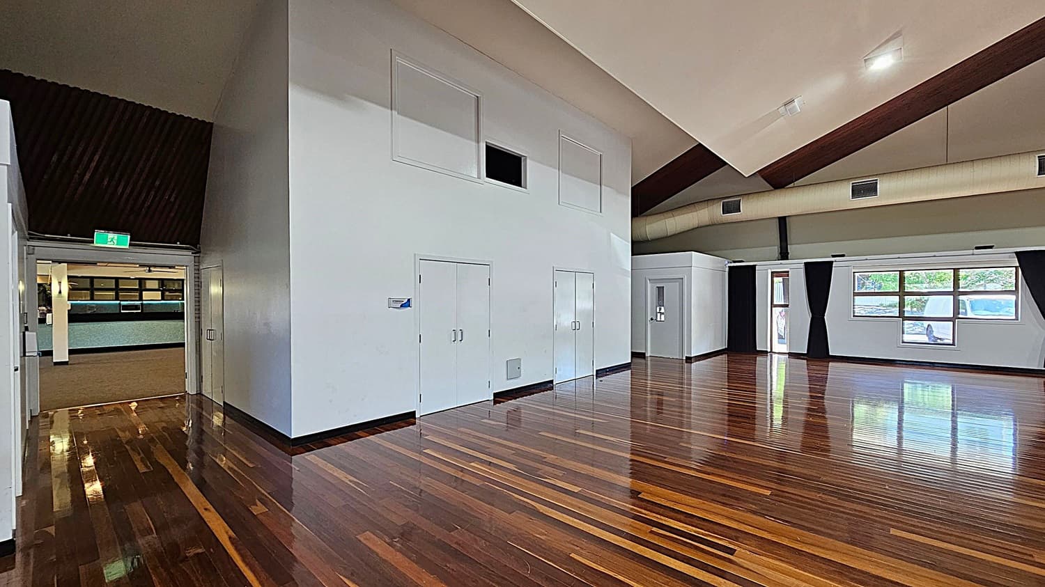 A spacious room with polished wooden floors and a high, angled ceiling. On the left, there's an open doorway leading to another area with a visible exit sign. The centre features a large white wall with two sets of double doors and a small rectangular opening above. On the right, there are windows with black curtains, allowing natural light to enter. The ceiling has exposed beams and ventilation ducts.