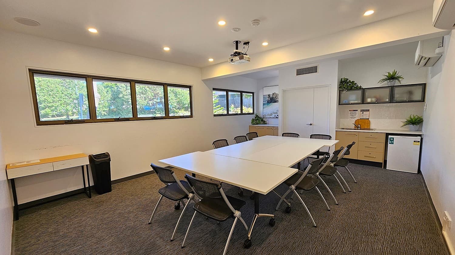 A hallway with a view into a meeting room. On the left, there's a counter with a metal urn and a "Stay COVID safe" sign. The floor is carpeted. On the right, a large potted plant stands next to a dark blue sofa. The meeting room has a table with chairs, cabinets, and plants on top. The walls are light-coloured, and there's a sign reading "Meeting Room 1" next to the door. Ceiling lights illuminate the space.