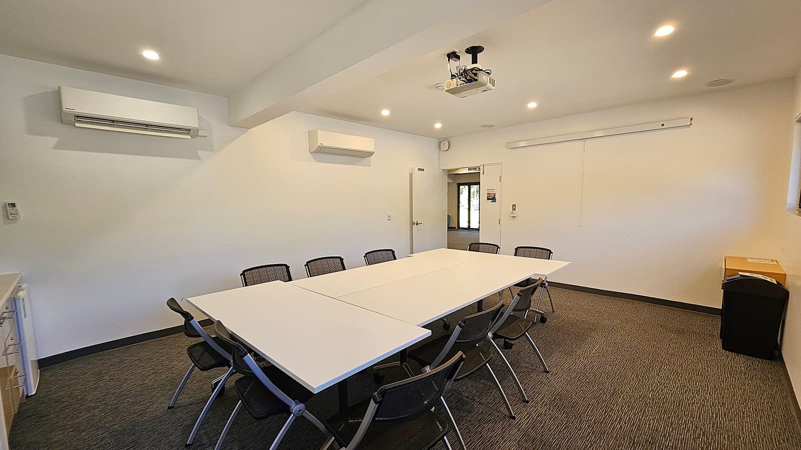 A conference room with a rectangular table in the centre surrounded by black mesh chairs. The walls are white, with two air conditioning units mounted on the left and back walls. A ceiling-mounted projector is above the table. The floor is carpeted in a green and grey pattern. On the right, there's a black bin and a cardboard box. The back wall has a door leading to another room, and a whiteboard is mounted on the wall. The ceiling has recessed lighting.