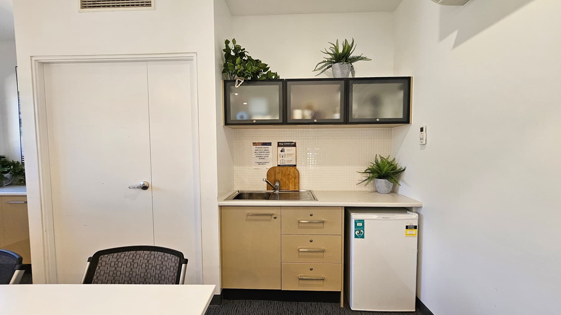 A small kitchenette with a white countertop and a stainless steel sink. Above the sink are three frosted glass cabinets with potted plants on top. Below the counter are beige cabinets and a mini fridge. A cutting board and a potted plant are on the counter. On the left, there's a closed white door with a silver handle. A black mesh chair is partially visible in the foreground. The floor is carpeted in dark grey.