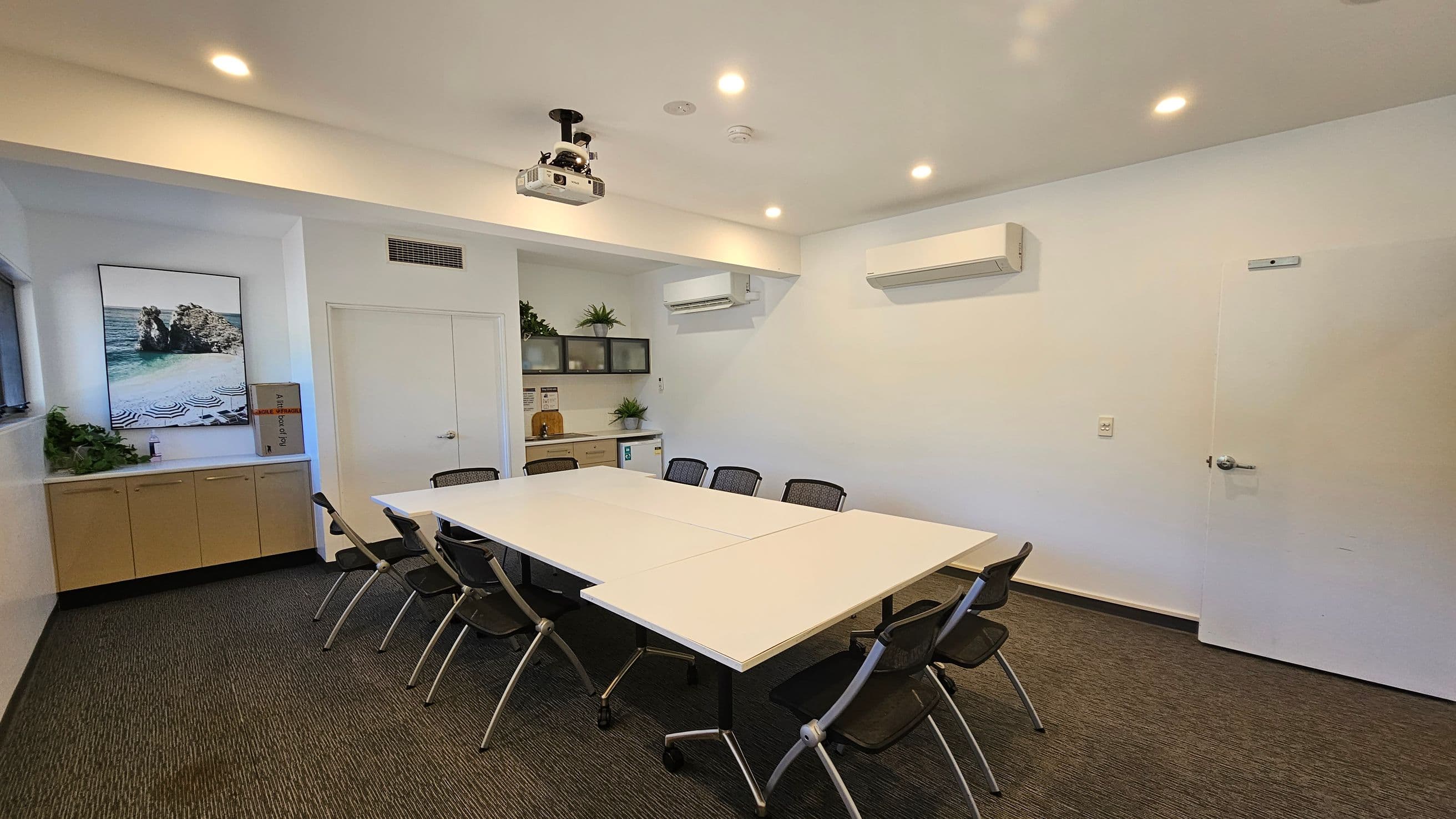 A conference room with a large rectangular table in the centre surrounded by black mesh chairs. On the left, a cabinet with plants and a framed picture of a coastal scene. The back wall has a door and a small kitchenette with more plants. Two air conditioning units are mounted on the walls. The ceiling is equipped with recessed lights and a projector. The floor is carpeted in a mix of grey and green tones.
