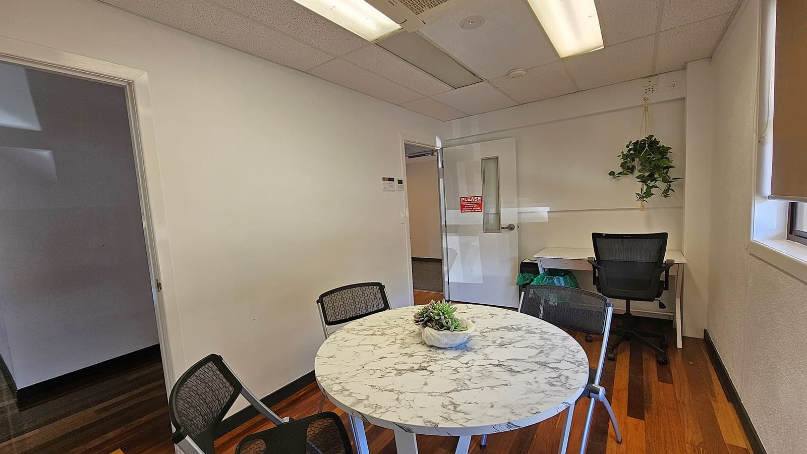 A small office room with a round marble-patterned table in the foreground, surrounded by three chairs. A potted plant sits on the table. In the background, there's a desk with a black office chair and a hanging plant above. The walls are white, and there's a door with a sign. The floor is wooden, and the ceiling has fluorescent lights. A window with a blind is on the right.
