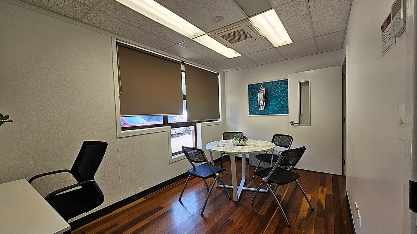 A small office room with wooden flooring and a white ceiling with fluorescent lights. On the right, there's a white door with a small window. In the centre, a round table with a decorative plant is surrounded by four black mesh chairs. A painting of a boat on water hangs on the wall. On the left, a window with brown blinds partially open reveals a glimpse of a red car outside. A black office chair is positioned near a white desk.