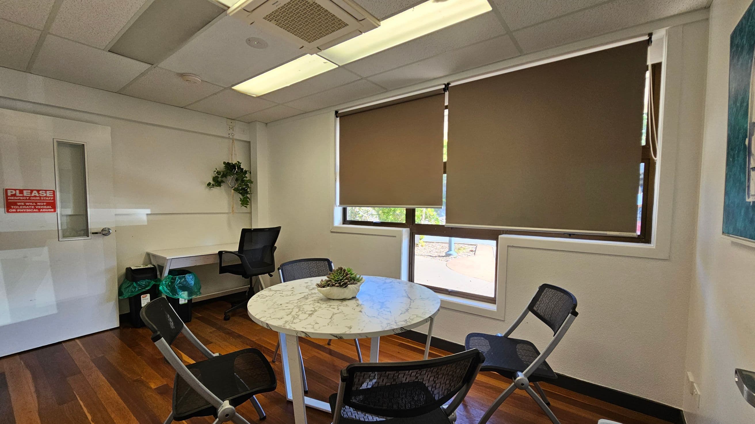 A small office room with a round marble table in the centre, surrounded by four black mesh chairs. On the right, a window with brown blinds partially drawn. A potted plant sits on the table. To the left, a white door with a red sign and a desk with a black office chair. Above, fluorescent lights are set in a drop ceiling. The floor is wooden with a polished finish. A small plant hangs on the wall near the desk.