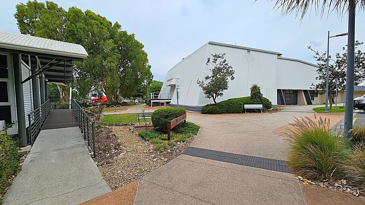 A modern building with a curved roof, identified as Coolum Beach Library, is in the centre. It has large windows and a ramp leading to the entrance. A signpost in the foreground indicates dogs must be on a leash. The surrounding area features well-maintained greenery and trees on both sides. The path in front is paved, with a small garden bed and a bicycle rack to the right.