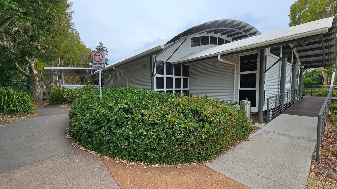 A modern building with a curved metal roof and large windows is on the right. A ramp leads up to the entrance. In the foreground, there is a dense green hedge. On the left, a path curves around the hedge, with a "Skateboards Prohibited" sign visible. Tall trees with thick trunks and green foliage are in the background, providing shade. The ground is a mix of concrete and tan-coloured paving.