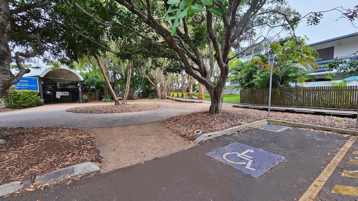 A pathway curves through a park-like area with trees and fallen leaves. On the left, there's a building with a sign and a covered entrance. To the right, a wooden fence and a building with balconies are visible. In the foreground, a parking space marked for disabled access is on the asphalt. A yellow barrier and a signpost are near the centre, with lush greenery throughout the scene.