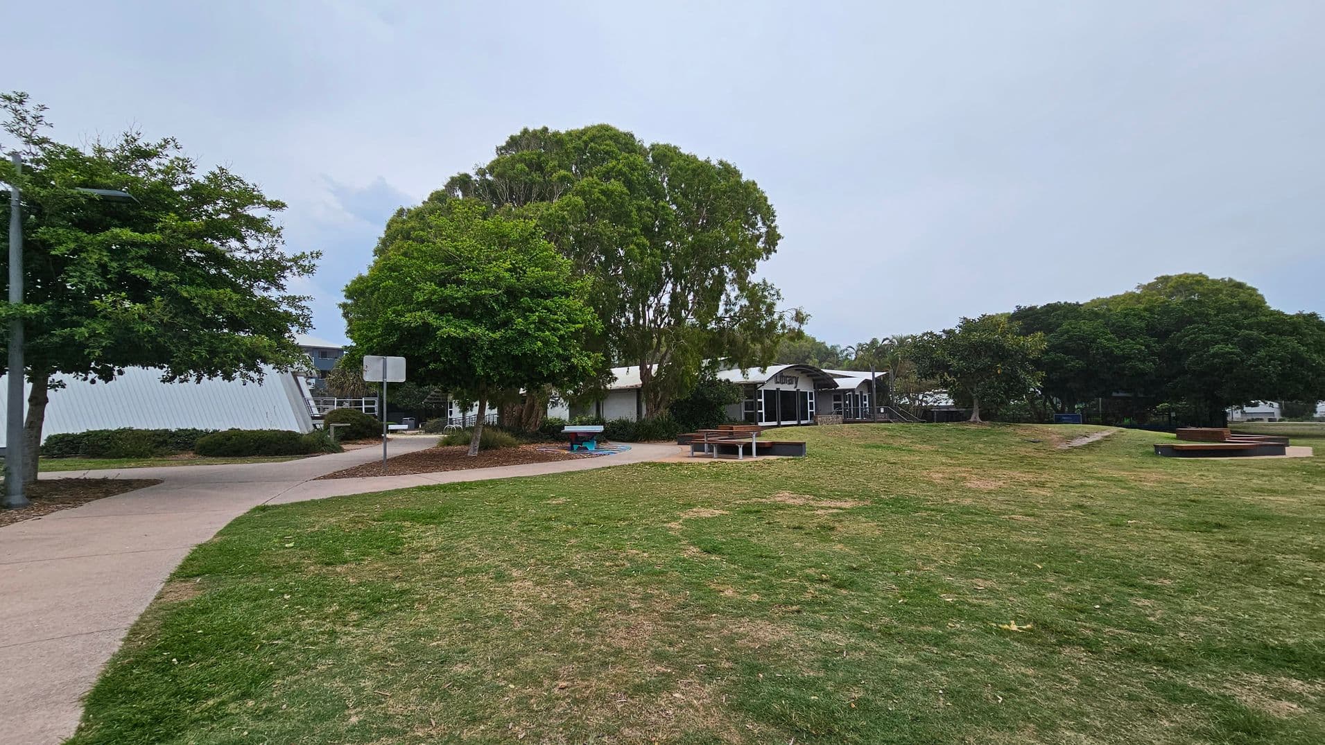 A grassy area with patches of brown is in the foreground. A paved path curves from the left to the centre, flanked by trees and bushes. In the middle, a building with a sign reading "Library" is partially obscured by large trees. On the right, there are benches and more trees. The background features additional greenery and a clear sky.