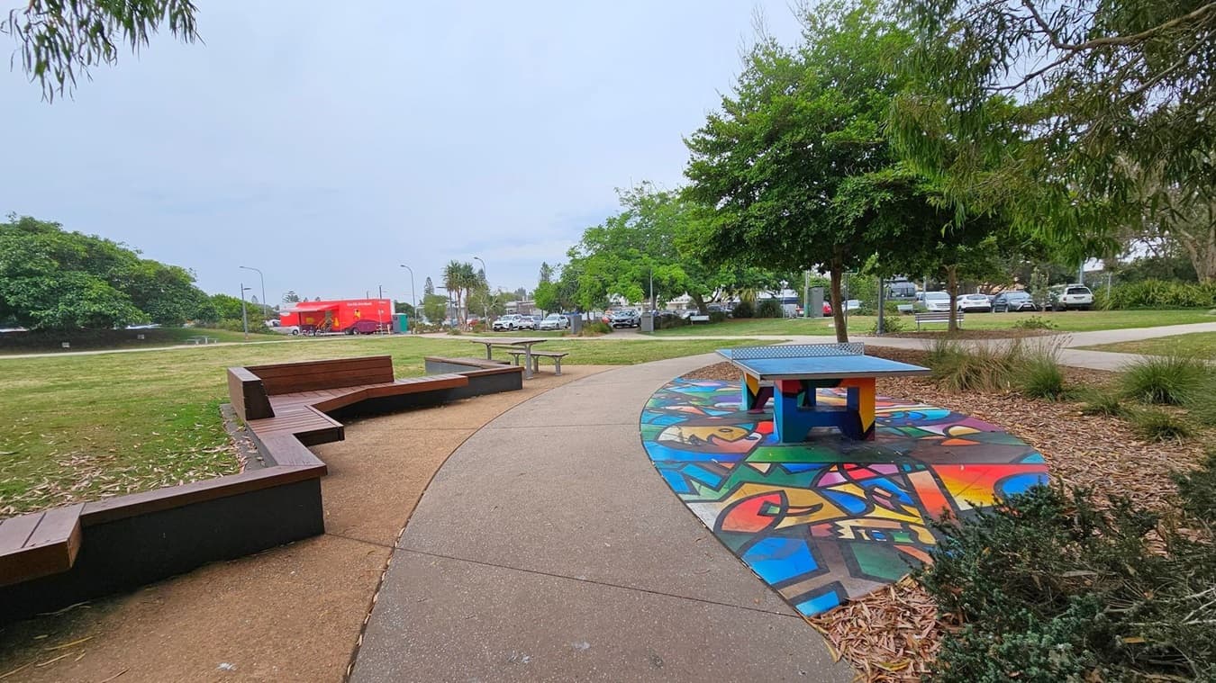 A park scene with a curved pathway in the centre. On the right, there's a colourful ping pong table on a vibrant painted surface. To the left, wooden benches form a seating area. In the background, a red food truck is parked on the grass, with several cars visible behind it. Trees and shrubs are scattered throughout the park, providing greenery. The ground is covered with a mix of grass and mulch.
