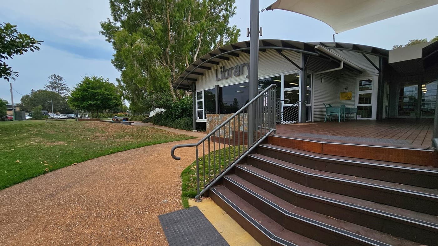 A library building with a curved roof is on the right, featuring a sign that reads "Library" above the entrance. There are steps with a metal railing leading up to the entrance, and a small seating area with green chairs. On the left, a paved path curves through a grassy area with trees and a few parked cars in the background. The area is surrounded by greenery, creating a peaceful setting.