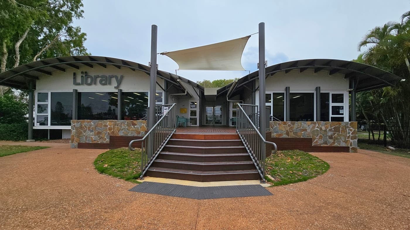 A modern library building with a curved roof and large windows. The entrance is in the centre, accessible by a set of stairs with metal railings. The facade features stonework and a sign reading "Library" on the left. A white shade sail is suspended above the entrance. The ground is a mix of gravel and grass, with trees visible on both sides.