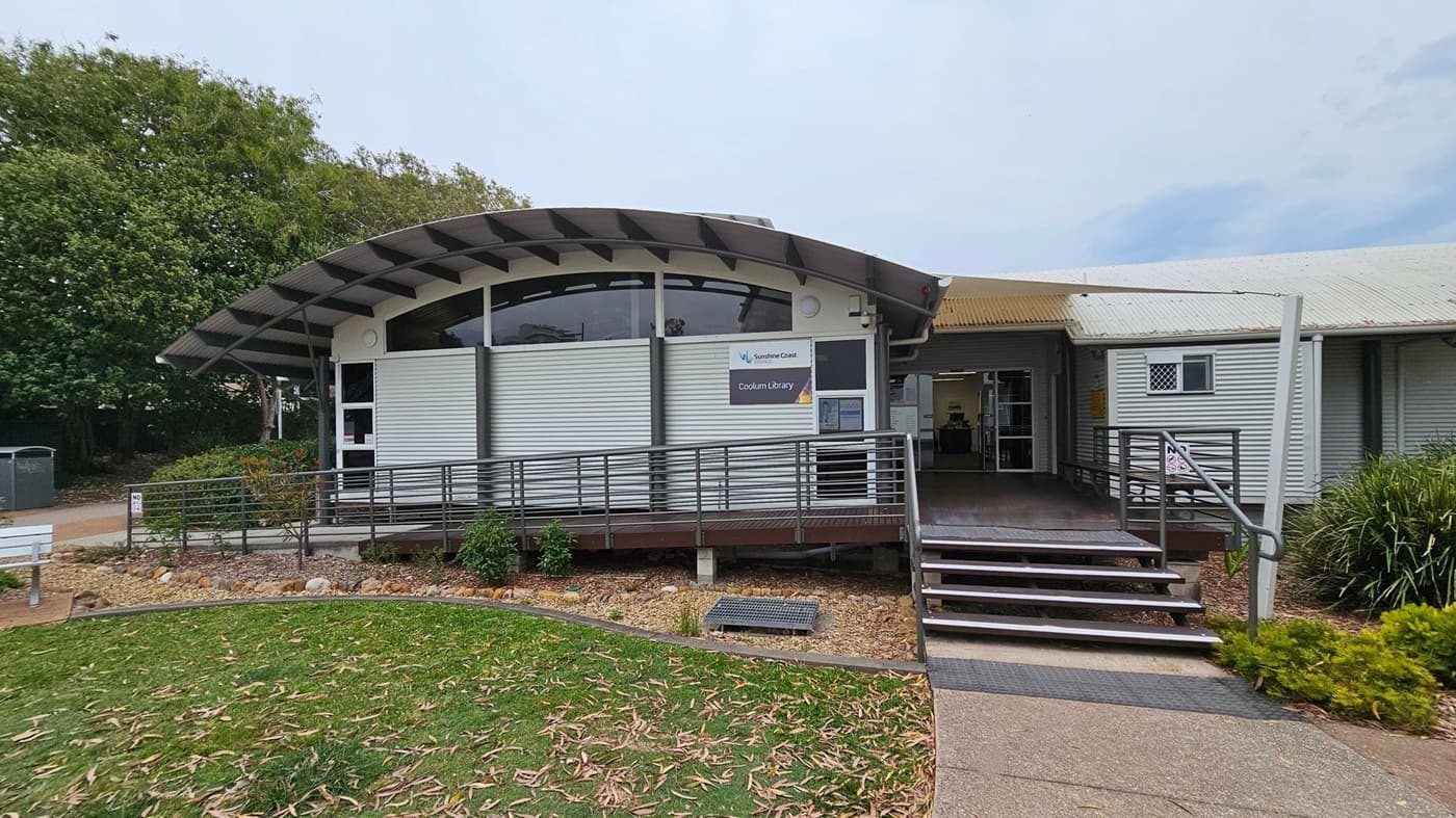 A modern building with a curved metal roof and a ramp leading to the entrance. The front has a sign reading "Coolum Library" and features large windows. The exterior is clad in corrugated metal. On the left, there are trees and a small garden area with a bench. The right side has a pathway bordered by bushes. The ground is a mix of grass and concrete, with some fallen leaves scattered around.