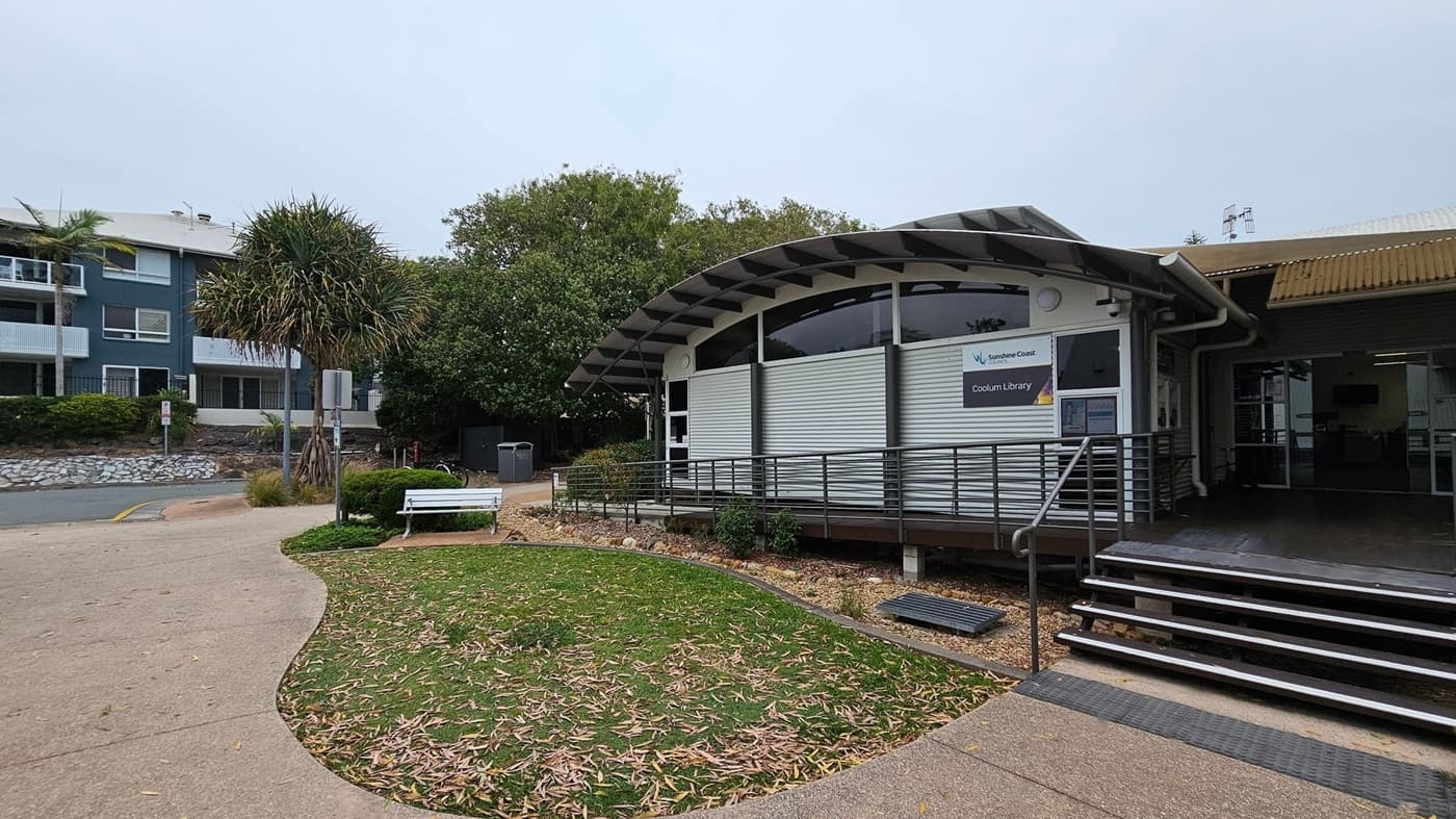 A modern building with a curved roof on the right, identified as Coolum Library. It has a metal ramp and steps leading to the entrance. In front, there's a small garden with grass and scattered leaves. A white bench and a signpost are on the left, near a paved path. In the background, there are trees and a multi-storey building with balconies. The ground is a mix of concrete and grass.
