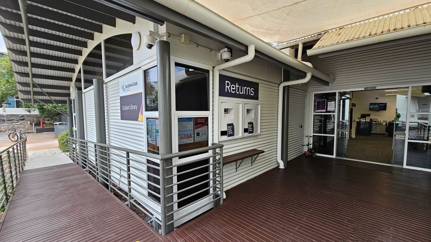 A library entrance with a corrugated metal exterior. On the right, there are two book return slots labelled "Returns" and a small shelf beneath them. The entrance has glass doors leading inside, where a person is visible. On the left, a wooden ramp with metal railings leads up to the entrance. Above, a corrugated metal awning provides cover. A bicycle is parked outside on the left, near some greenery. The floor is wooden decking.