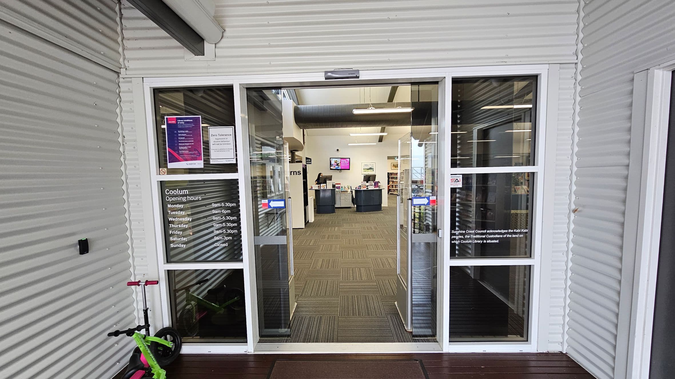 A glass entrance door with opening hours and notices displayed. To the left, a bright green and pink scooter is parked against the corrugated metal wall. Inside, a reception desk is visible with a person standing behind it. Bookshelves and a digital screen are in the background. The floor is carpeted with a striped pattern, and the ceiling has exposed ducts and lights.