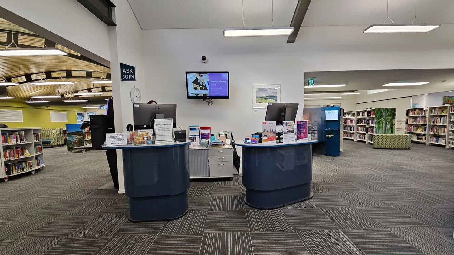 A library interior with a reception desk in the centre, featuring two computer monitors and various informational pamphlets. A person stands behind the desk on the right. The floor is covered with grey and green patterned carpet tiles. To the left, there are bookshelves and a curved wooden ceiling with hanging lights. In the background, more bookshelves and a green patterned seating area are visible. A wall-mounted screen and a clock are on the wall behind the desk.