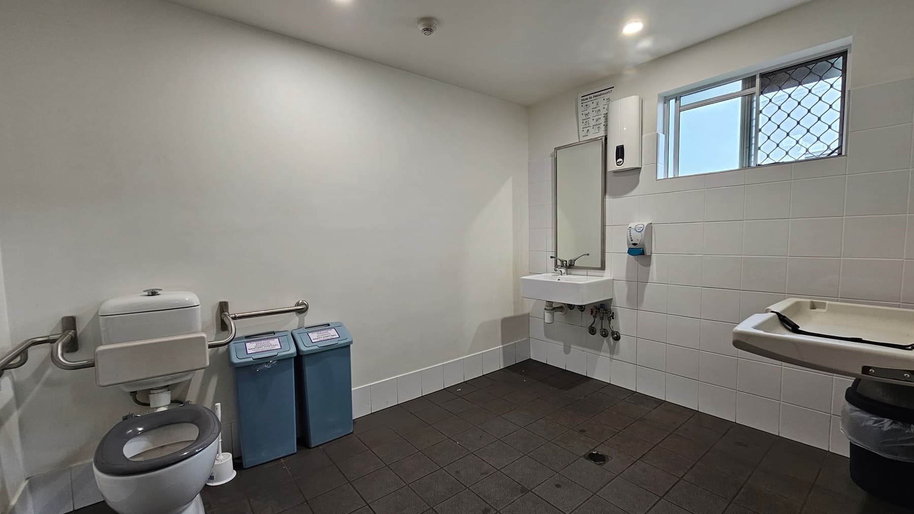 A public restroom with a grey-tiled floor and white walls. On the left, there is a toilet with support rails and two blue bins beside it. In the centre, a sink with a mirror above it is mounted on the wall. To the right, a baby changing station is attached to the wall. Above the sink, a small window with a security grille allows natural light in. The ceiling is white with recessed lighting.