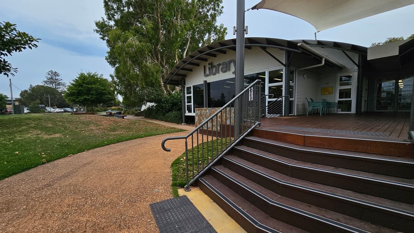 A library building with a curved roof is on the right, featuring a sign that reads "Library" above the entrance. There are steps leading up to the entrance with a metal railing. A small outdoor seating area with green chairs is visible. On the left, a path curves through a grassy area with trees and a few parked cars in the background. The ground is a mix of grass and a textured path.