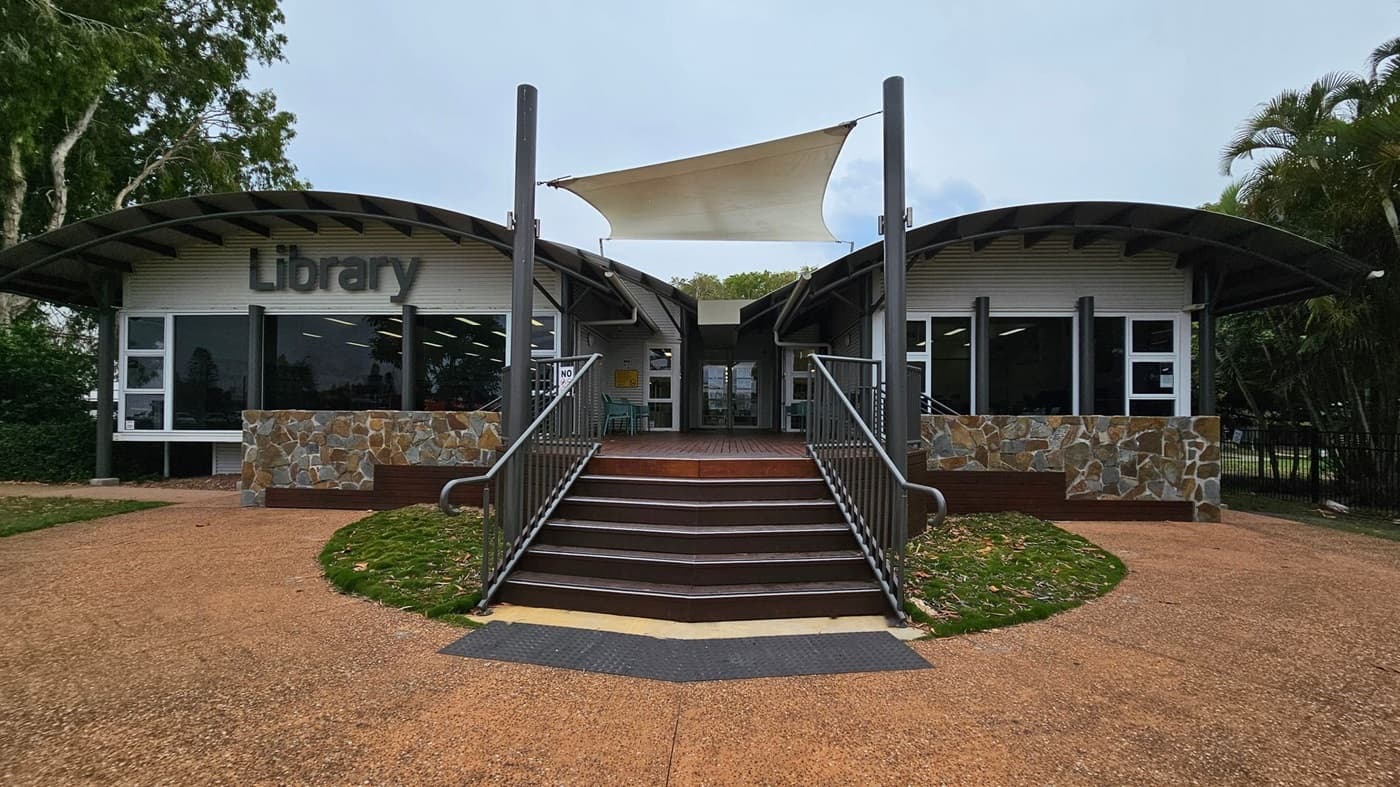A modern library building with a curved roof and large windows. The entrance is in the centre, accessible by a set of stairs with metal railings. The facade features stonework and the word "Library" in large letters on the left side. A white shade sail is suspended above the entrance. The surrounding area includes trees and greenery, with a paved path leading to the entrance.