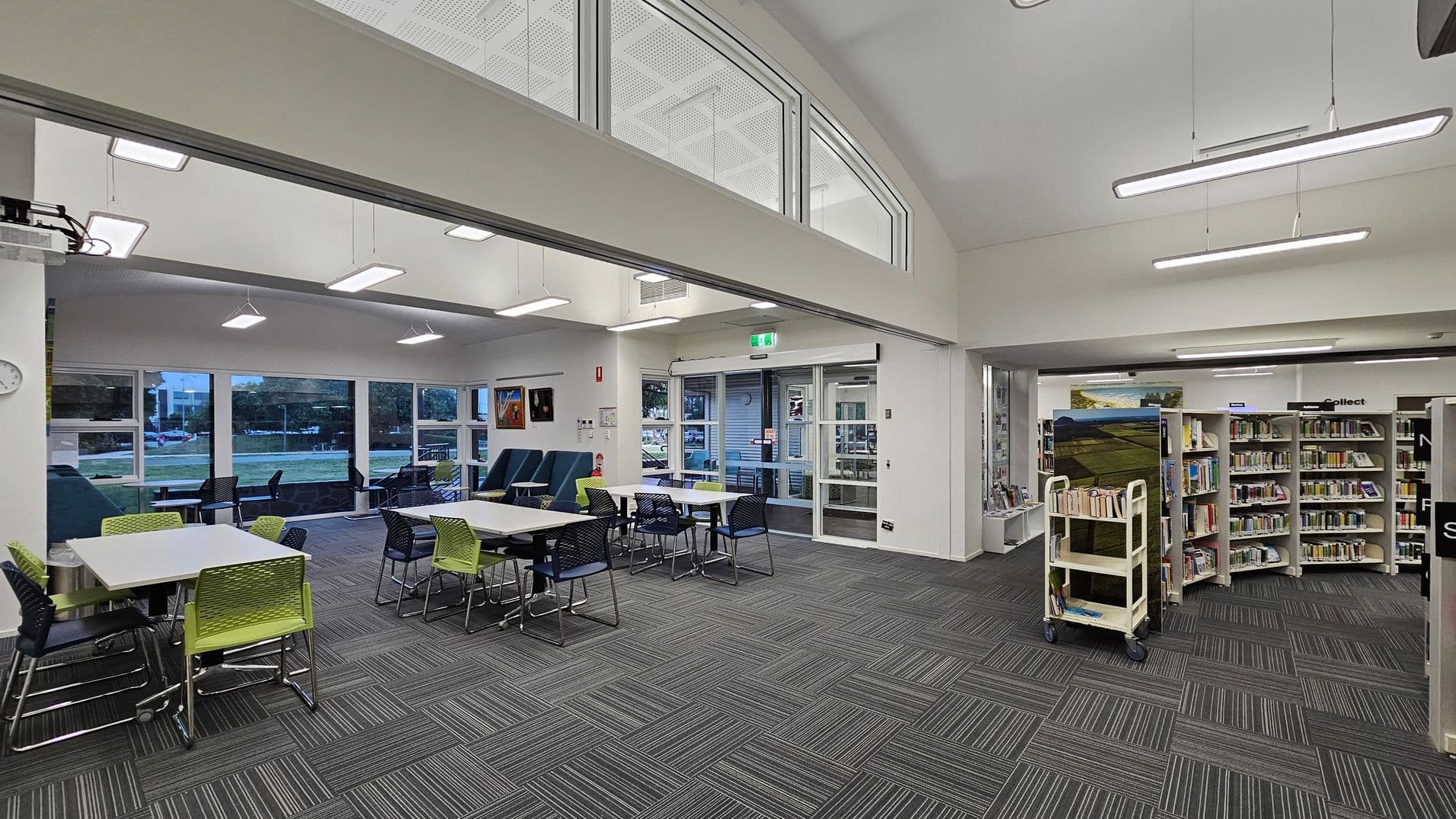 A modern room with a grey carpeted floor and a white perforated ceiling. In the centre, there are tables surrounded by green and black chairs. On the left, a banner with "first 5 forever" and directional words. The right side features large glass doors leading to an outdoor area. The back wall has windows and two green sofas. Fluorescent lights hang from the ceiling, and a clock is mounted on the left wall.