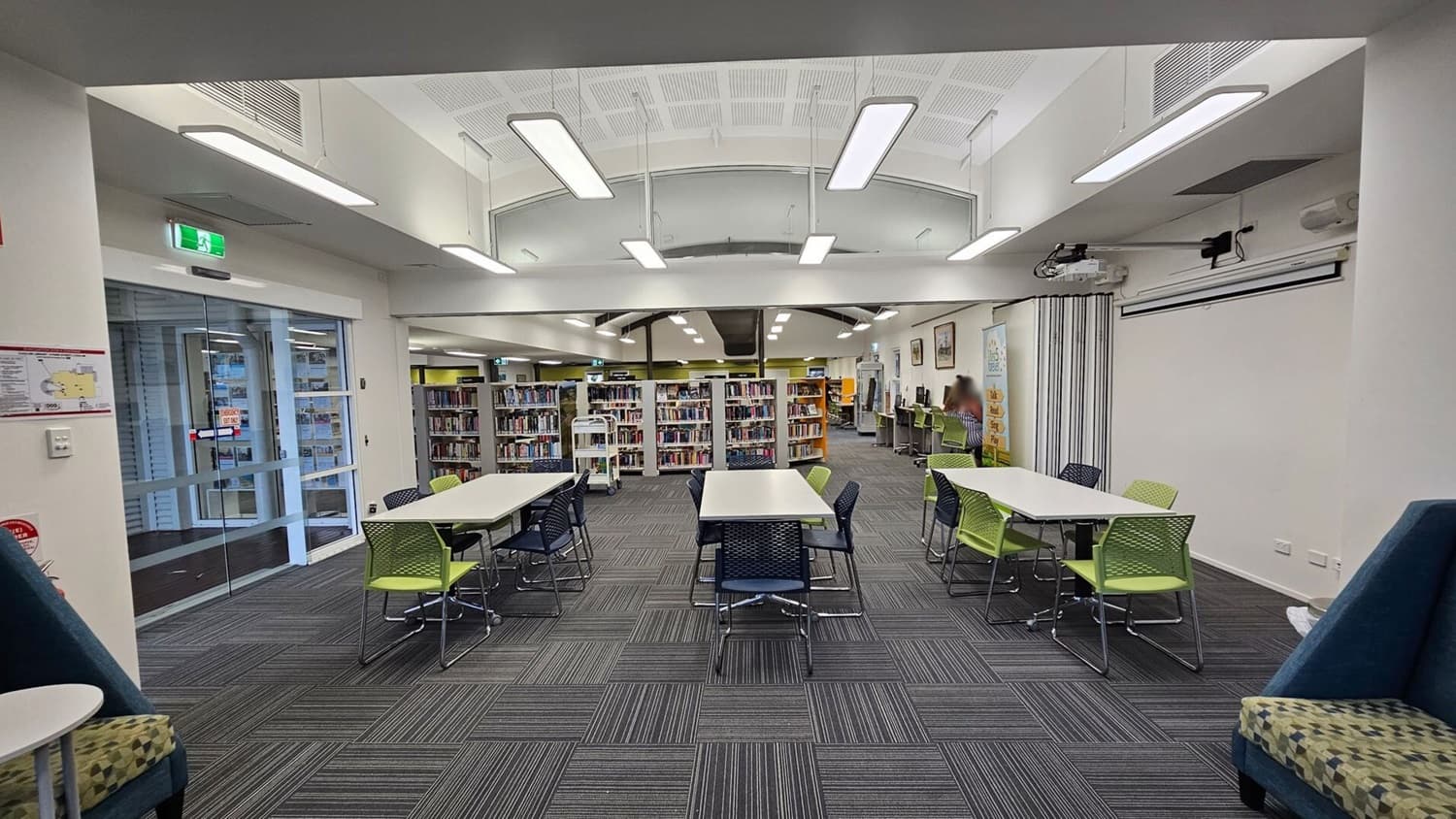 A library interior with grey carpeted flooring and a white ceiling featuring rectangular lights. In the foreground, there are tables with green and blue chairs. The background shows bookshelves filled with books. On the right, a person is seated at a computer desk. On the left, there is a glass door with an exit sign above it. The walls are light-coloured, and there are some framed pictures and a banner on the right wall.