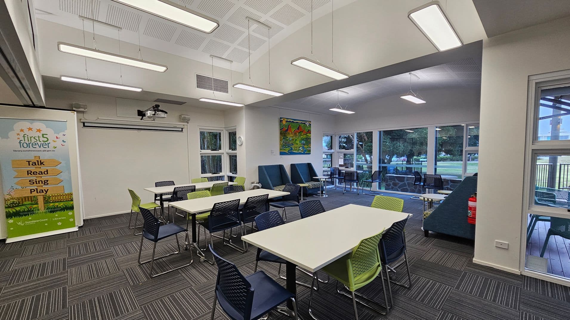 A modern library room with a grey and green striped carpet. On the left, a banner reads "first 5 forever" with activities listed. Several tables with green and black chairs are arranged in the centre. The back wall features a colourful painting and large windows showing a grassy outdoor area. Blue cushioned seating is near the windows. The ceiling has rectangular lights, and a projector is mounted above. A fire extinguisher is visible on the right.