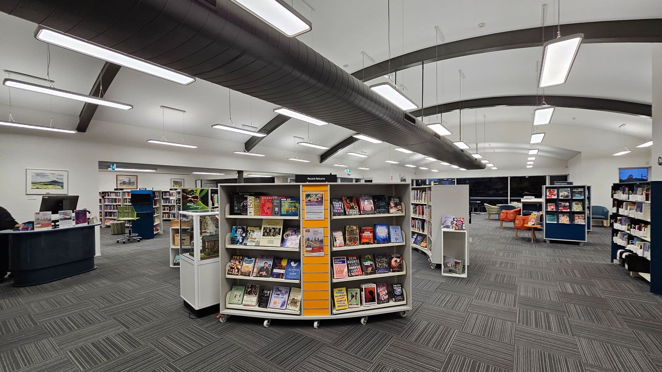 A library interior with a grey carpeted floor and a ceiling featuring fluorescent lights. On the right, there's a "Returns" section with black shelves. Nearby, a desk with a computer and a sign that reads "Ask Join" is visible. In the centre, there are white display shelves with books, some decorated with a pumpkin face. The left side has a wall with bookshelves filled with books, a door, and a seating area with a blue chair. The walls are painted yellow with a colourful border.