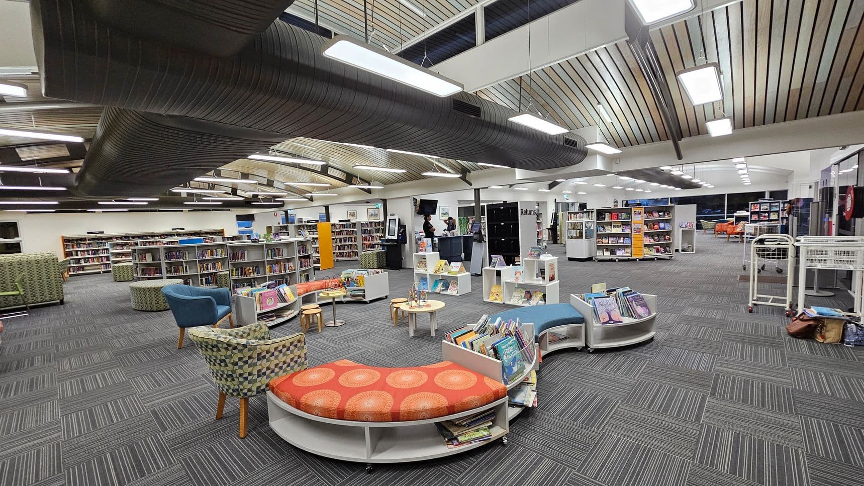 A modern library interior with grey carpeted flooring and a wooden panelled ceiling. In the foreground, there are curved bookshelves filled with children's books and colourful seating, including a blue chair and a patterned armchair. The middle area has more bookshelves and a small table. In the background, a counter with people standing and more bookshelves are visible. Overhead, large black air ducts and bright rectangular lights are suspended from the ceiling.