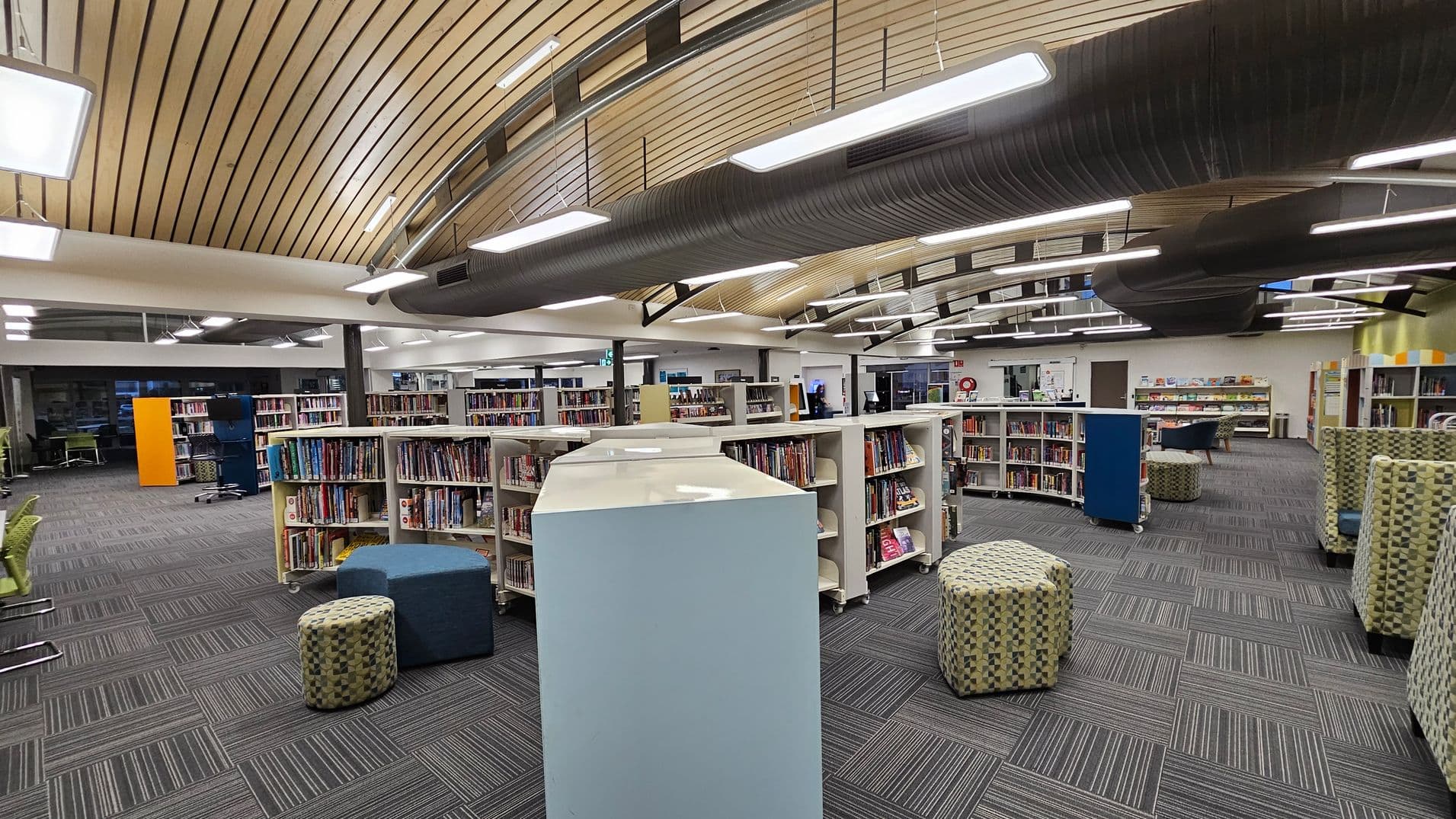 A modern library interior with a curved wooden ceiling and exposed black ductwork. The floor is covered with grey carpet tiles featuring a striped pattern. In the centre, there are white bookshelves filled with books, and several patterned ottomans and a blue pouffe for seating. On the left, green chairs are visible, and on the right, more seating and bookshelves. Bright overhead lighting illuminates the space.