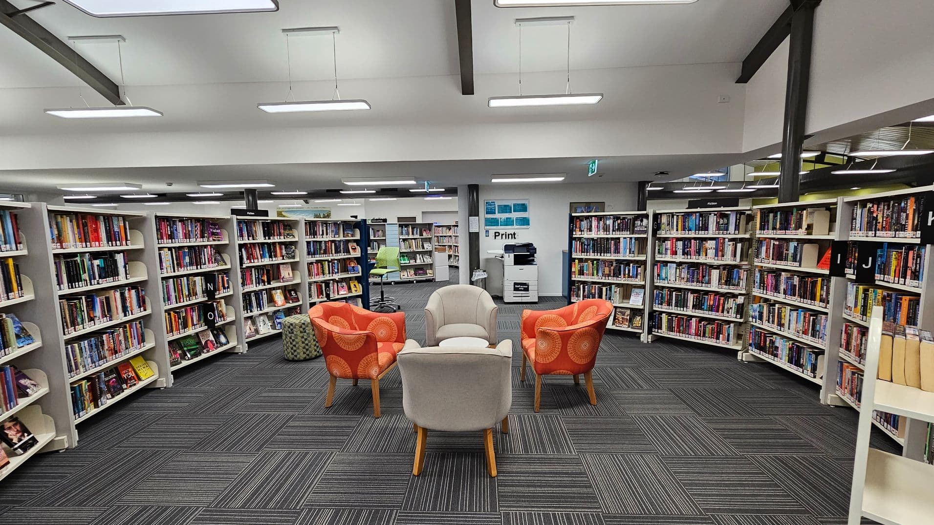 A library interior with bookshelves lining the walls on the left and right, filled with various books. In the centre, there is a seating area with a mix of orange and beige armchairs arranged in a circle on a grey carpet with a striped pattern. The ceiling is white with modern rectangular lights, and there is a printer station against the back wall.