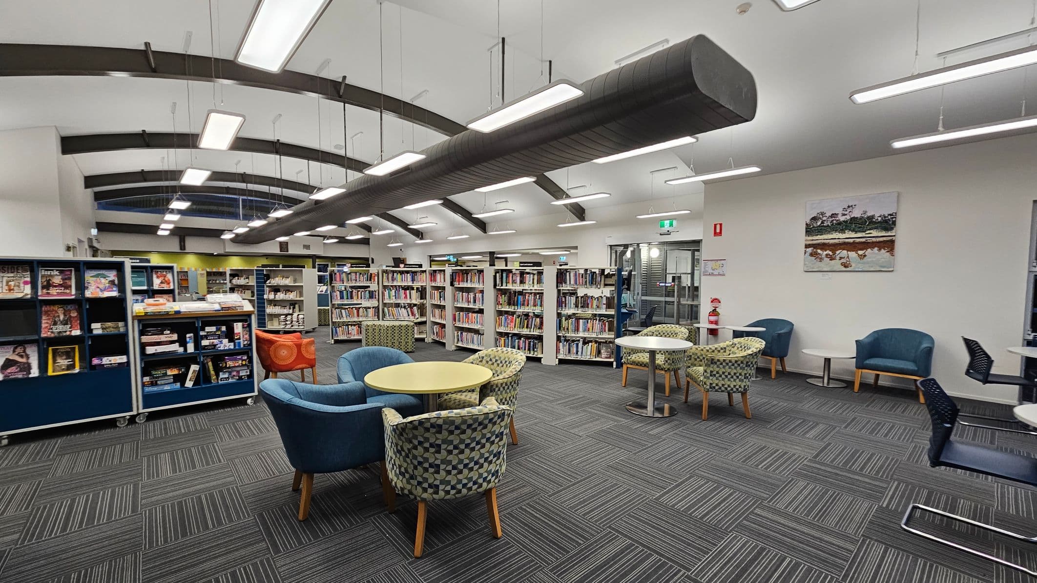A library interior with a modern design. On the left, there are shelves filled with books and magazines. In the centre, there are round tables surrounded by patterned and solid-coloured chairs. The right side features more seating with small tables and a fire extinguisher nearby. The ceiling has exposed beams and hanging lights, and the floor is covered with dark, striped carpet tiles. A painting hangs on the wall in the background.