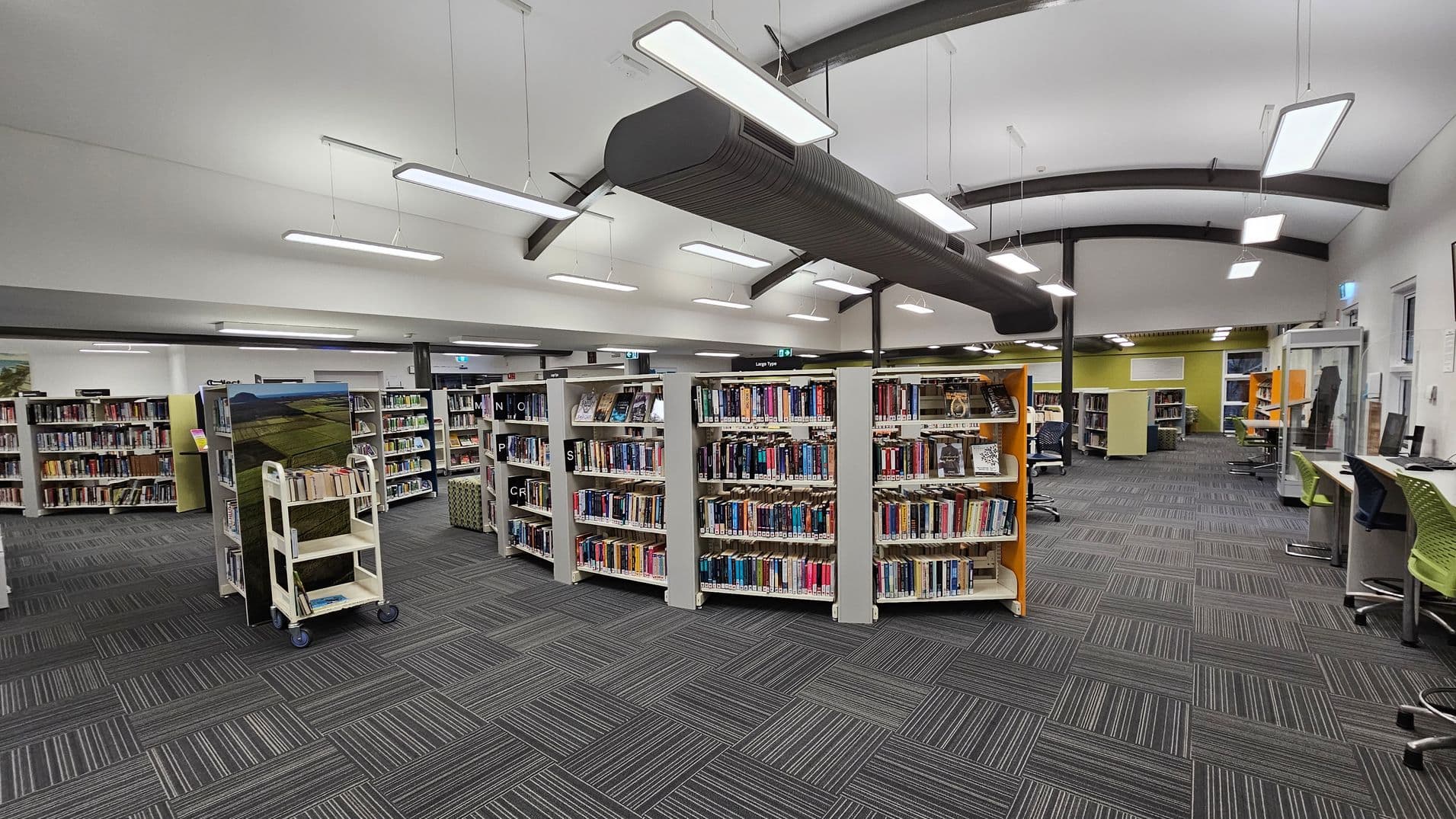 A modern library interior with grey carpeted flooring featuring a striped pattern. In the centre, there are several bookshelves filled with books. On the left, a book trolley is visible. To the right, there are computer workstations with green chairs. The ceiling is white with exposed beams and hanging rectangular lights. In the background, more bookshelves and a green wall are visible.