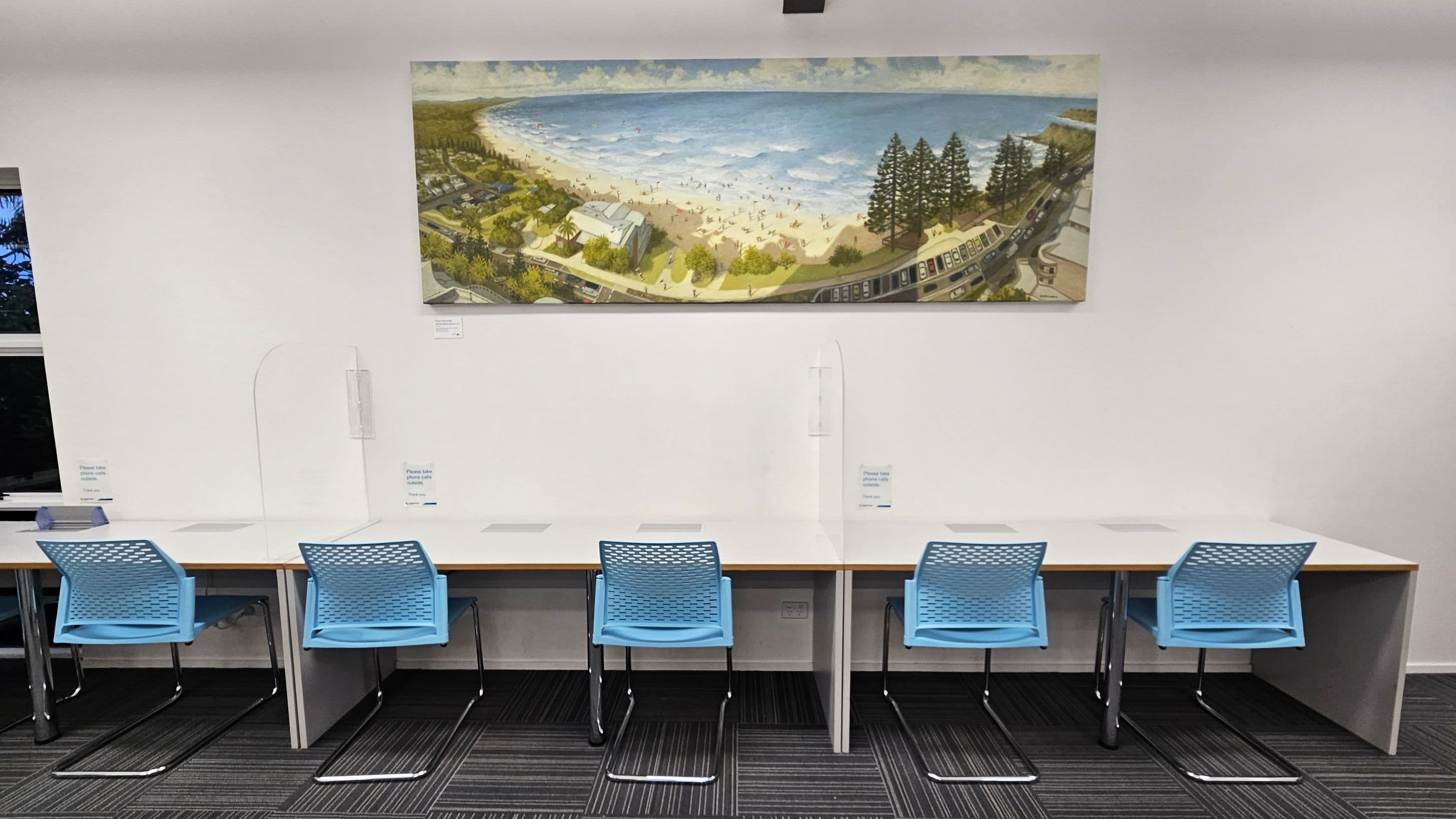 A row of four blue chairs is positioned in front of a long white desk with partitions, creating individual study spaces. Above the desk hangs a large painting depicting a beach scene with people, trees, and buildings. The floor is covered with dark striped carpet tiles. The wall is plain white, and there is a window on the left side.