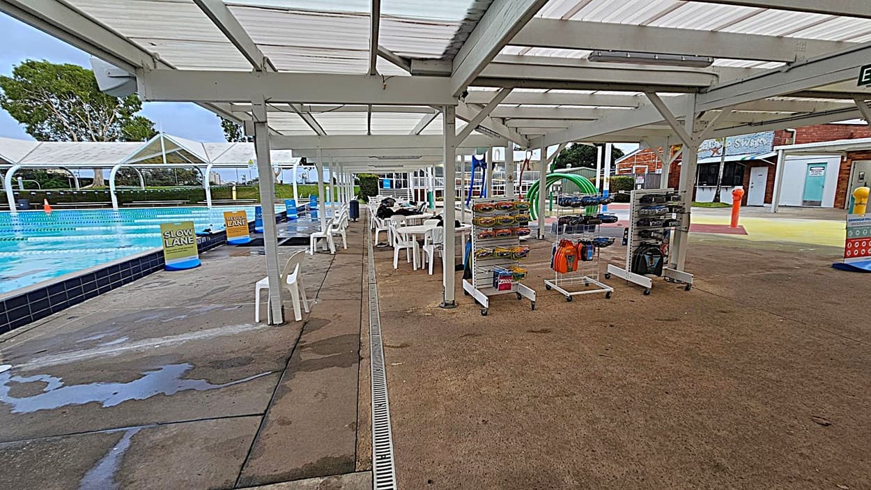 Outdoor swimming pool area with a covered walkway. On the left, a pool with lane markers and signs for "Slow Lane" and "Medium Lane". White plastic chairs and tables are under the shelter. On the right, racks with swimming gear and accessories. The ground is concrete, and the ceiling is a white, slatted structure. In the background, there are brick buildings and a grassy area with trees.