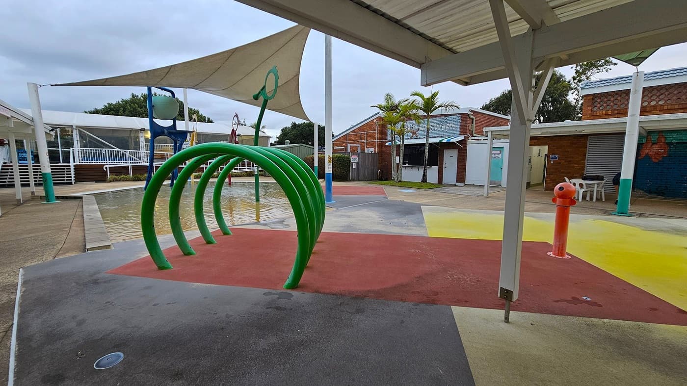 A playground with a splash pad area featuring a series of green arches on the left. The ground is covered with colourful rubber surfaces in red, yellow, and green. In the background, there are brick buildings with blue roofs and a few palm trees. A white shade sail is overhead, and a red water feature stands on the right. The area is surrounded by white railings and a small pool of water.