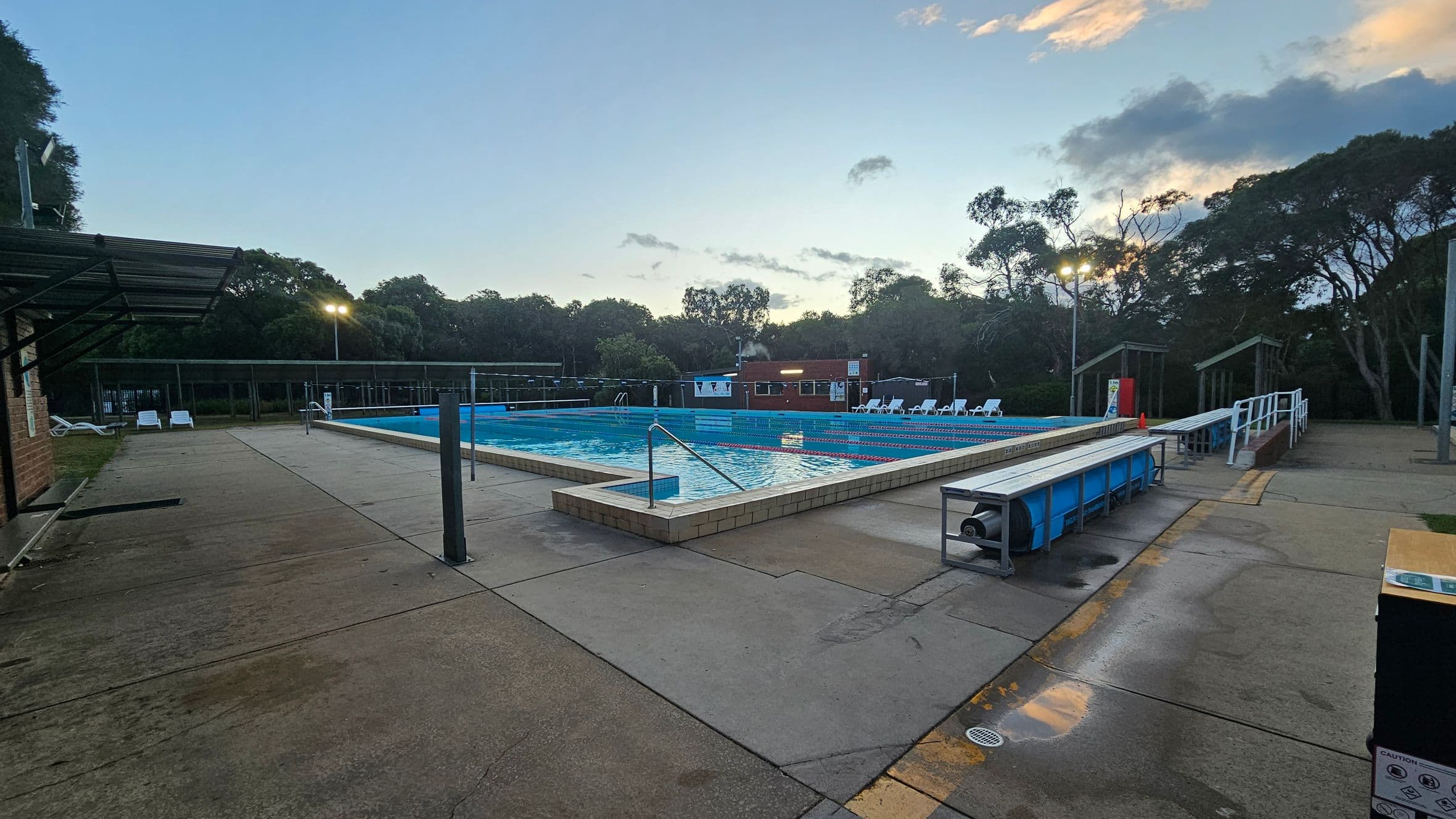 Outdoor swimming pool surrounded by a concrete deck. On the left, there is a covered area with white lounge chairs. The pool is in the centre, with lane dividers visible. On the right, more lounge chairs are lined up, and a blue pool cover roller is nearby. Trees and a small building are in the background. The area is lit by tall lights, and the sky has some clouds with a hint of sunset colours.