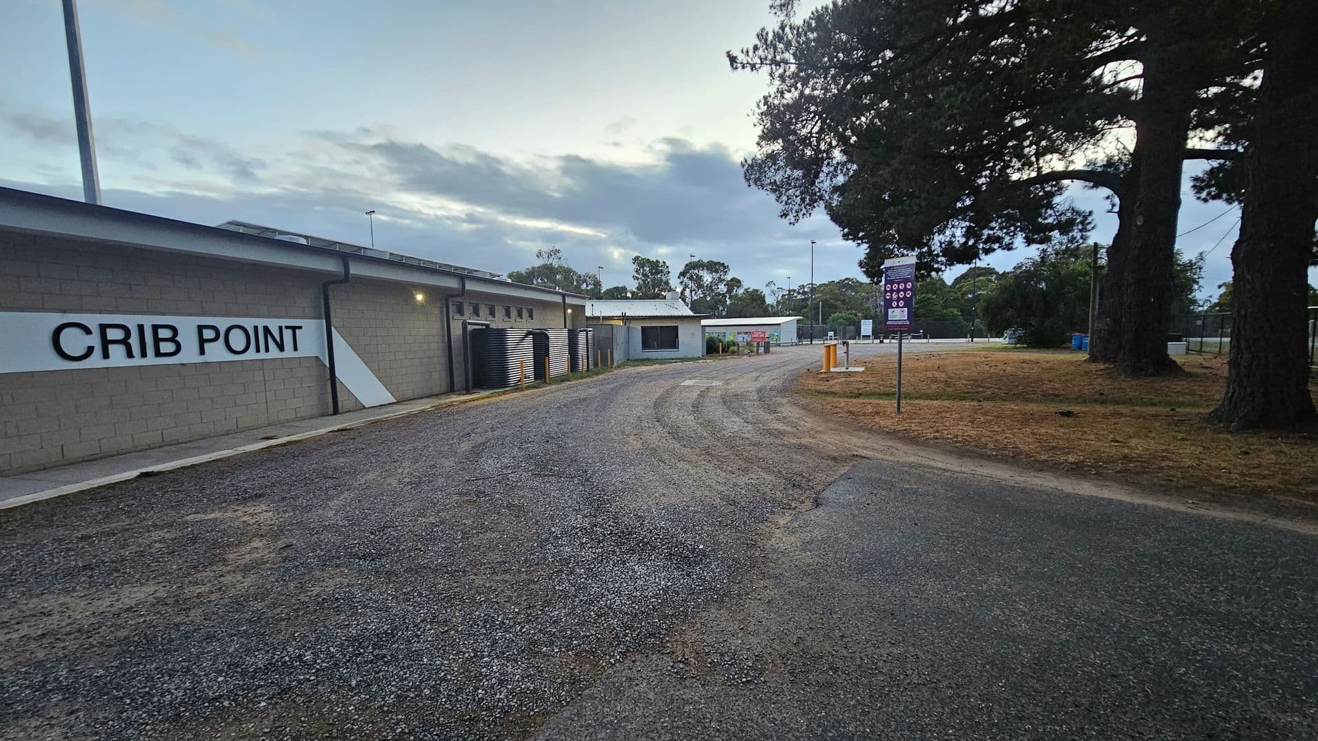 A gravel path curves through an outdoor area. On the left, a building with a sign reading "CRIB POINT" is visible, with several black water tanks lined up against it. On the right, there are large trees and a grassy area with a signpost. In the background, more buildings and trees can be seen, along with a few streetlights. The sky is overcast, casting a muted light over the scene.