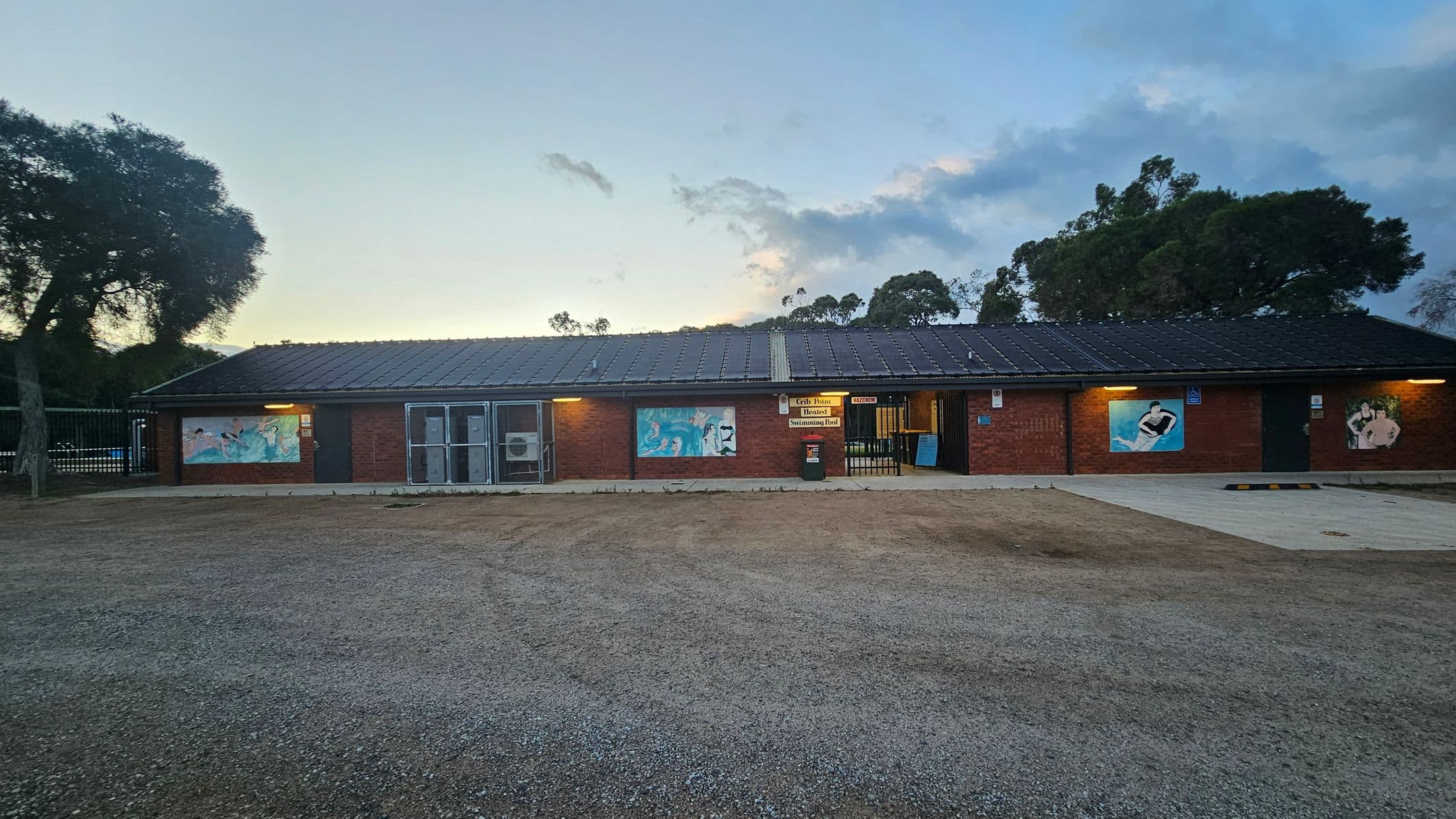 A single-storey brick building with a dark roof, featuring three large murals on its exterior walls. The entrance is centrally located, with a sign above it. The ground in front is a mix of gravel and sparse grass. Trees are visible in the background, and the sky is overcast. The building is lit by exterior lights, casting a warm glow on the walls.
