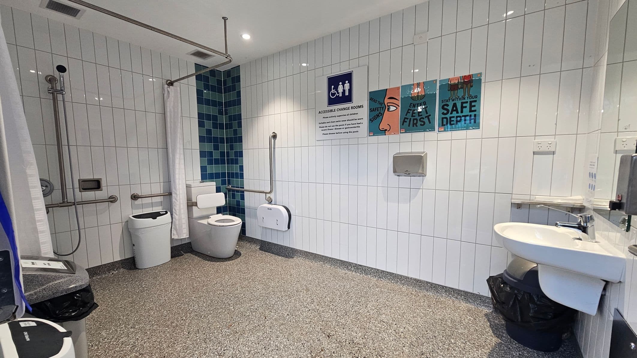Accessible bathroom with white tiled walls and a speckled floor. On the left, a shower area with a curtain, grab bars, and a showerhead. Next to it, a toilet with support rails and a nappy bin. The right side features a sink with a mirror above and a soap dispenser. The wall displays signs about safety and accessibility. The ceiling is white with recessed lighting.