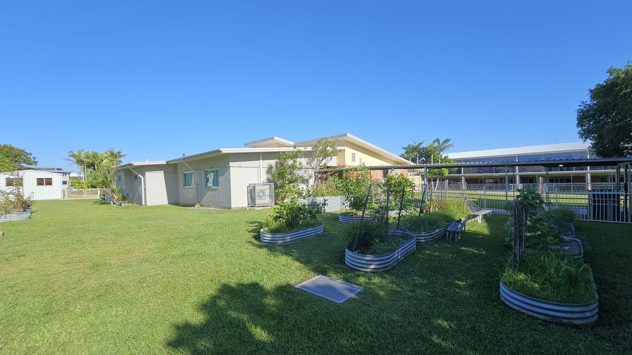 A grassy area with raised garden beds filled with plants is in the foreground. On the left, a light-coloured building with a sloped roof is visible, surrounded by a few small trees and shrubs. In the background, there is a larger building with a covered walkway and a metal fence. The sky is clear and blue.