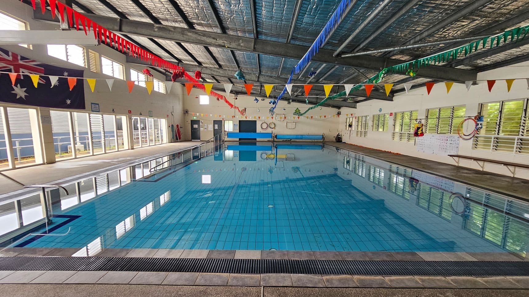 Indoor swimming pool with clear blue water, surrounded by a tiled deck. The ceiling is metal with exposed beams, adorned with colourful triangular bunting. On the left, large windows with louvres and an Australian flag. On the right, more windows and a wall with life rings and a notice board. The pool has a metal handrail on the left side. The floor is tiled, and the ceiling has insulation visible.