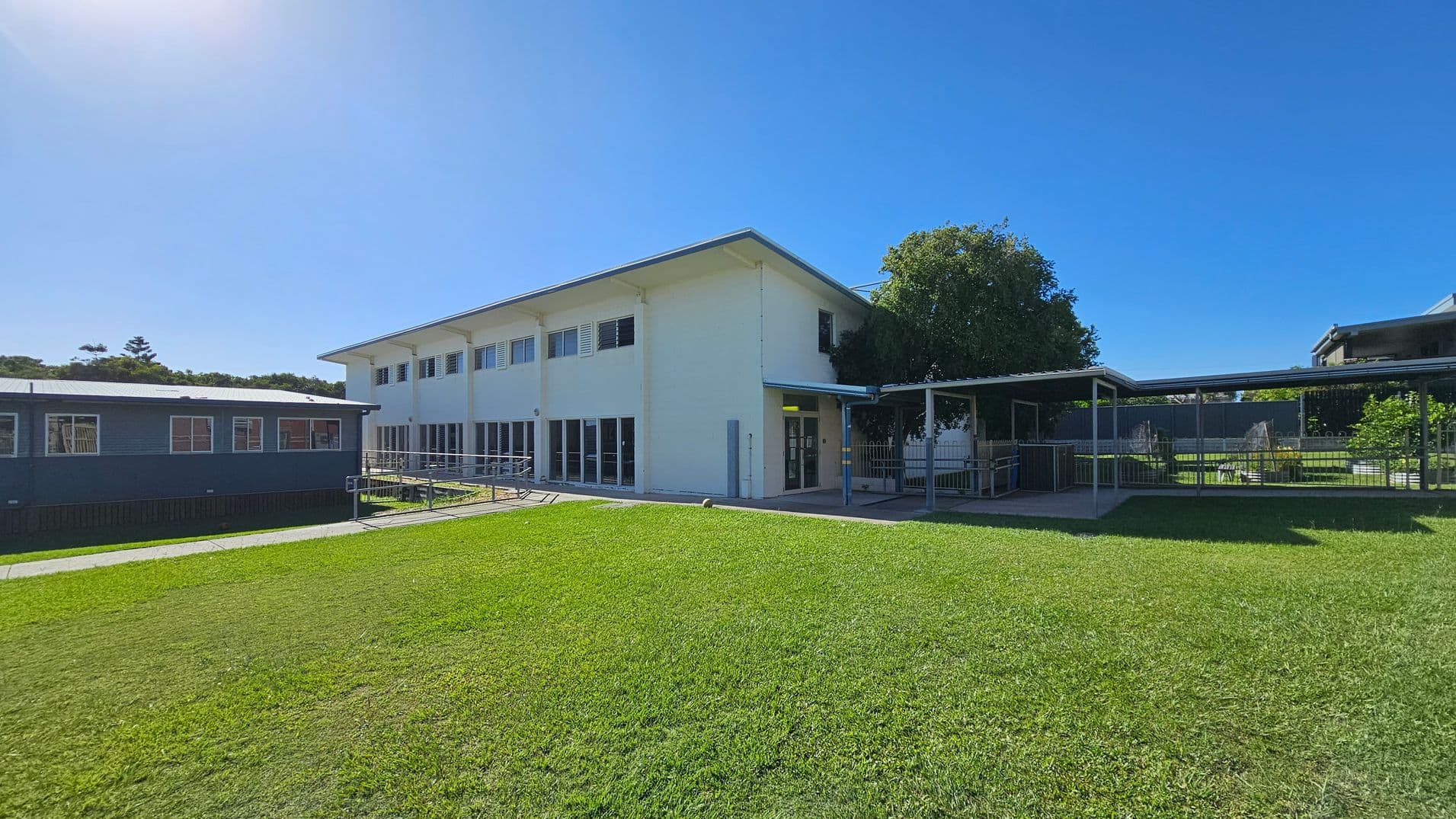 A two-storey white building with large windows on the right, connected to a blue single-storey structure on the left. A covered walkway extends from the main building, leading to a fenced area with greenery. The foreground features a well-maintained grassy lawn. A tree partially shades the entrance area. The sky is clear and blue.