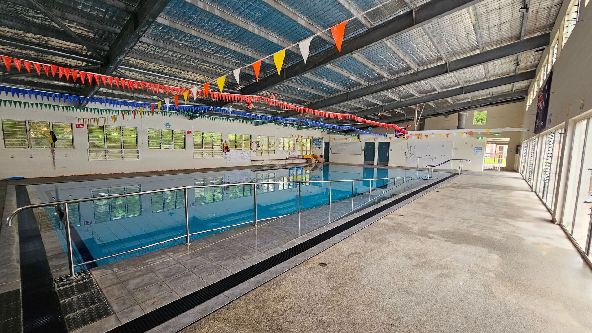 Indoor swimming pool with a tiled floor surrounding it. A metal railing runs along the pool's edge. The ceiling is made of exposed metal beams with colourful triangular flags hanging across. On the left, windows line the wall, allowing natural light in. The right side features large glass doors leading outside. The pool area is spacious, with a mix of tiled and carpeted flooring.