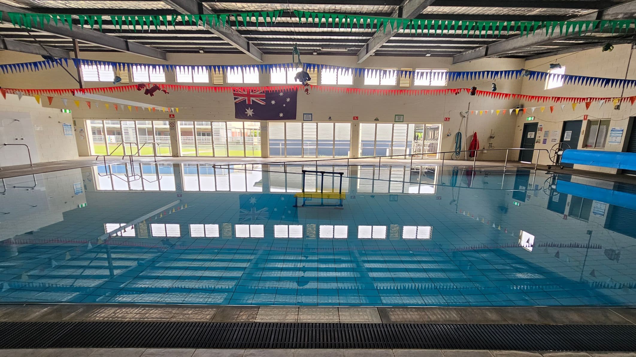 An indoor swimming pool with clear blue water. The ceiling has exposed beams and is adorned with colourful triangular bunting in green, blue, red, and yellow. Large windows line the far wall, allowing natural light to illuminate the space. An Australian flag is displayed on the wall. Pool equipment, such as a blue cover roller and a lifebuoy, is visible on the right. The floor around the pool is tiled, and a metal handrail is present at the pool's edge.