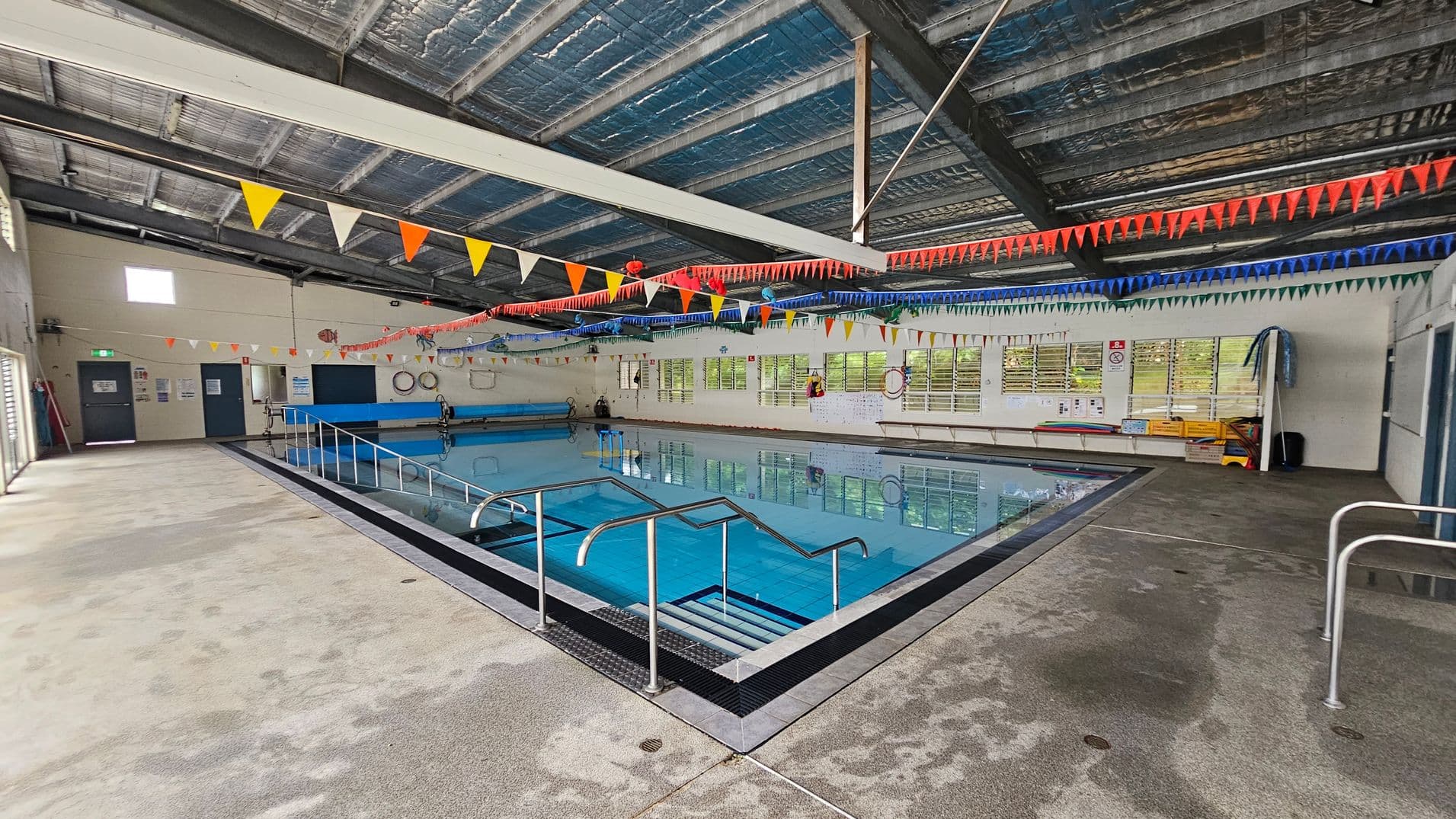 Indoor swimming pool with clear blue water, surrounded by a wet, textured floor. Metal handrails lead into the pool. Colourful triangular bunting hangs across the ceiling, which is metallic and industrial. On the left, a wall with doors and safety signs. On the right, windows with blinds, life rings, and pool equipment. The ceiling is high with exposed beams.