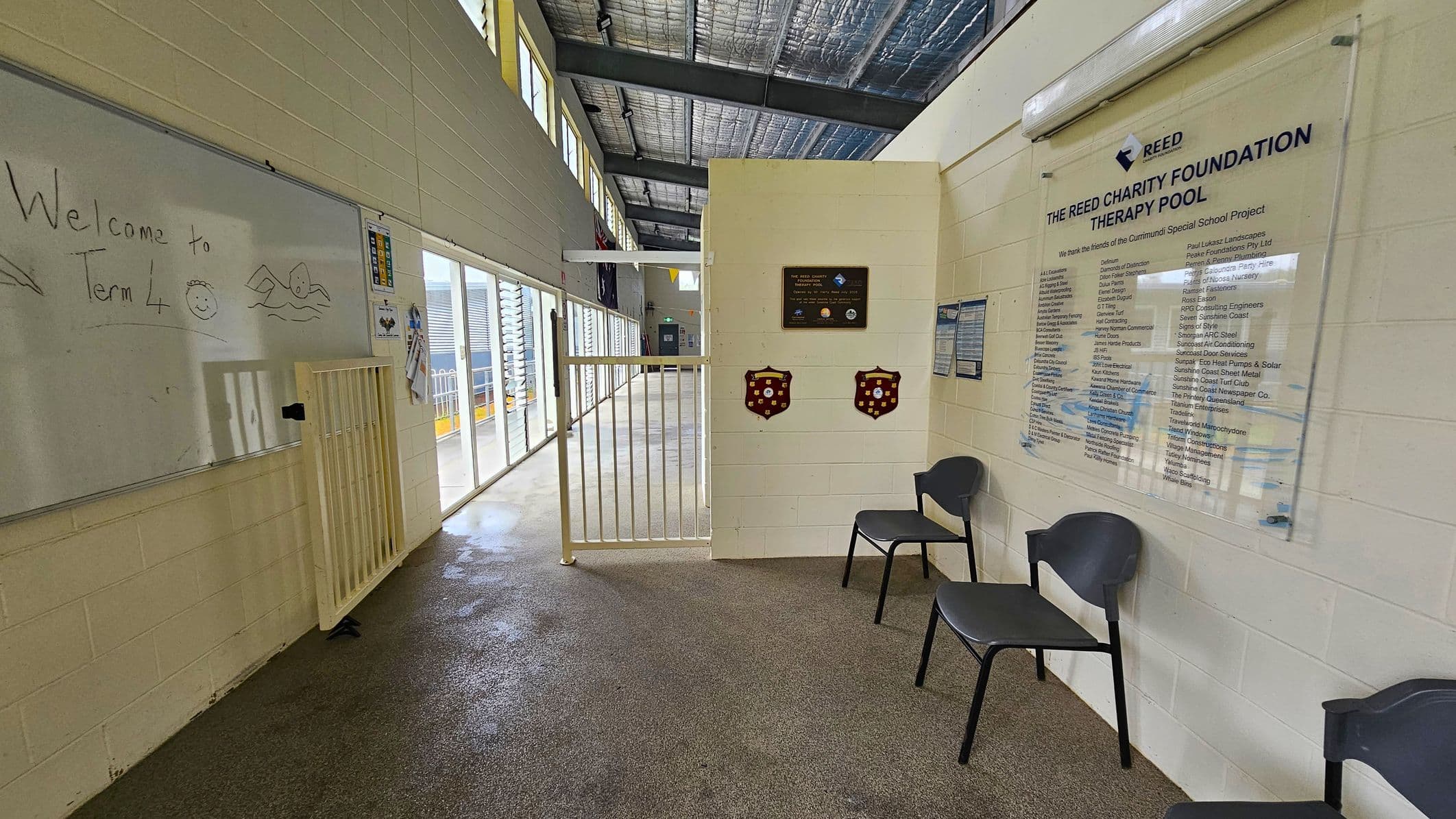 A hallway with a whiteboard on the left displaying "Welcome to Term 4" and a drawing of a swimmer. Below the board is a small gate. The right side features a wall with a plaque and a large sign for "The Reed Charity Foundation Therapy Pool," listing various names. Two black chairs are placed against the wall. The floor is a mix of wet and dry areas, and the ceiling has exposed metal beams and insulation. Large windows on the left allow natural light to enter.