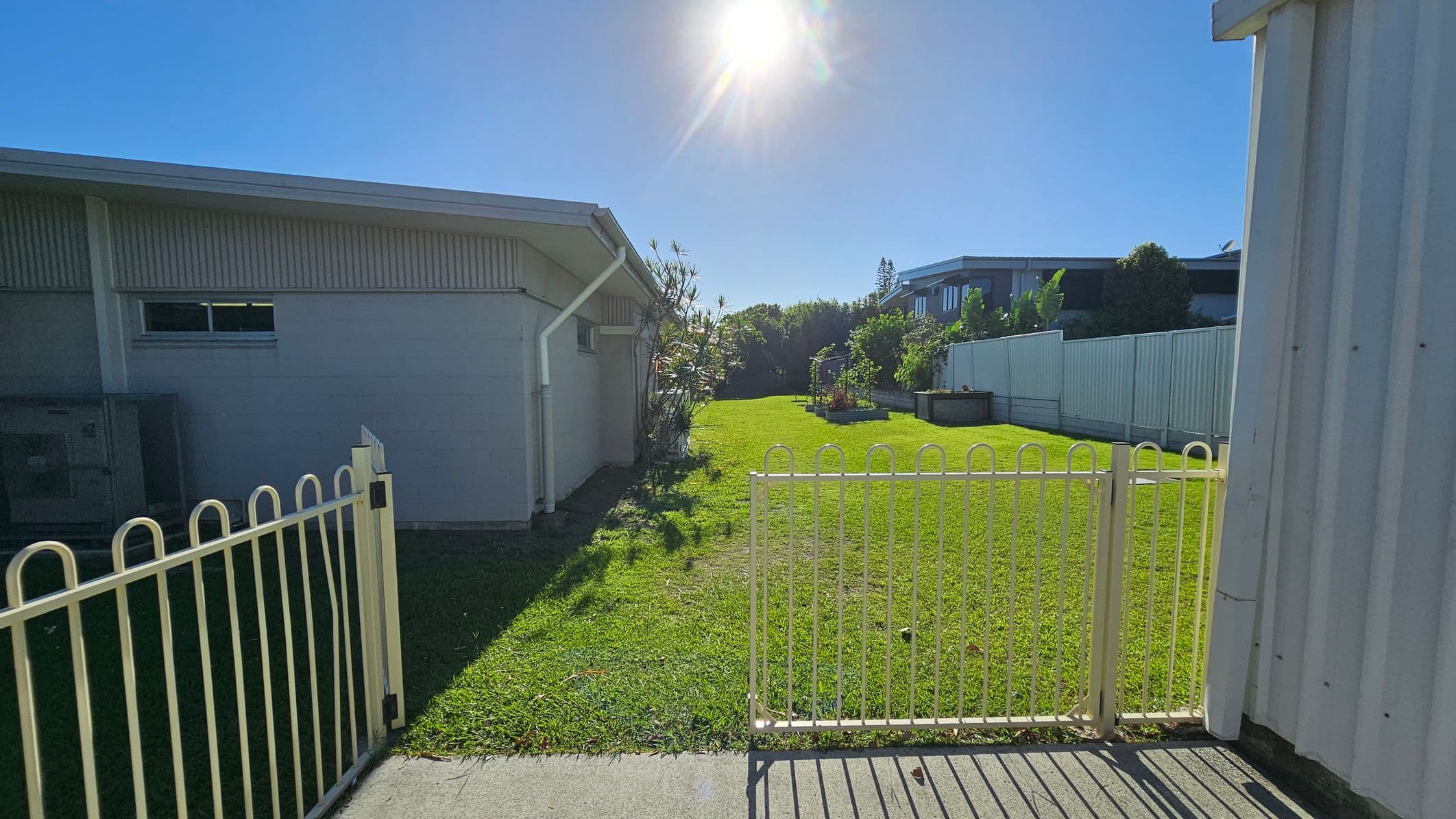 A grassy yard is bordered by a white fence, with a gate in the foreground. On the left, a building with a sloped roof and a small window is visible. On the right, another building with a corrugated exterior is partially seen. In the background, there are raised garden beds and a line of trees. The sun is shining brightly overhead, casting shadows on the grass.