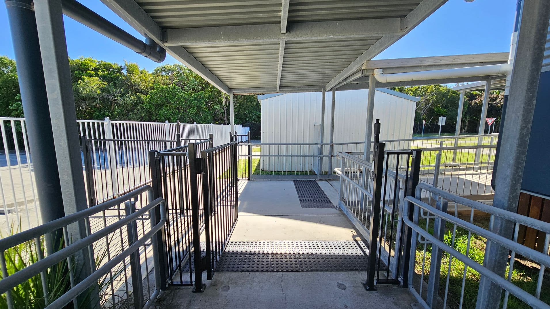 A covered walkway with a metal roof and railings on both sides. The floor is concrete with a textured mat in the centre. Two black gates are positioned along the path. In the background, there is a white building and a grassy area with trees. A metal fence runs parallel to the walkway on the left, and a yield sign is visible in the distance.