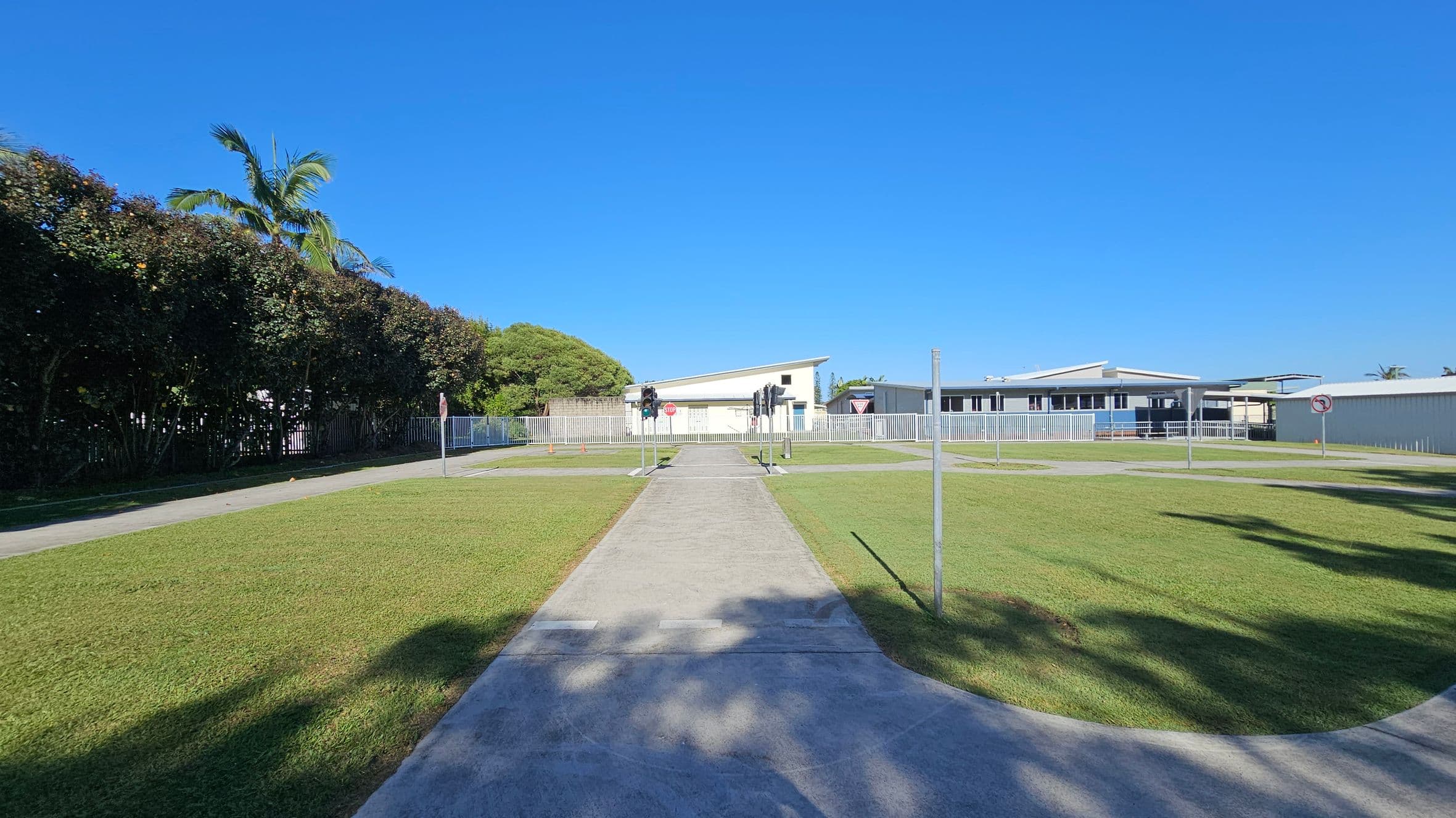 A paved path runs through the centre of a grassy area, leading to a building with a slanted roof in the background. On either side of the path are small traffic lights and signs, including a stop sign and a yield sign. To the left, a tall hedge with a palm tree is visible, while to the right, there are more buildings and a "no right turn" sign. Shadows from nearby trees are cast on the grass and path.